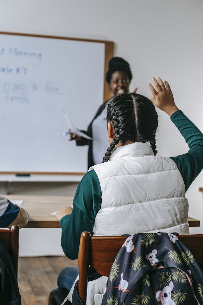 Smiling African American female teacher standing near whiteboard and looking at schoolgirl raising hand